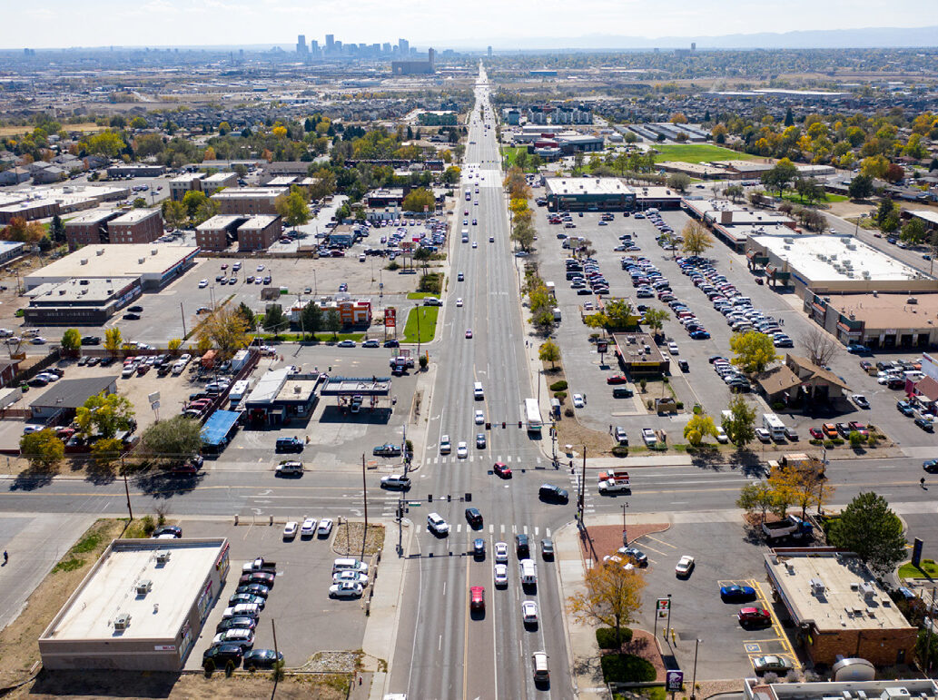 Aerial view of a wide road leading to a distant city skyline, with parking lots, buildings, and vehicles along the sides