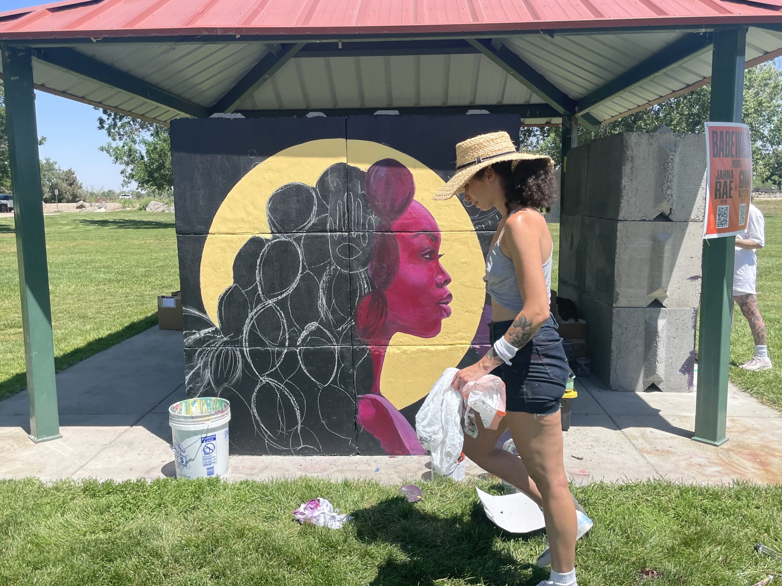 Woman painting a vibrant mural of a pink-faced person under a park shelter. Sign reads: BABEWALL now Johanna Rae.