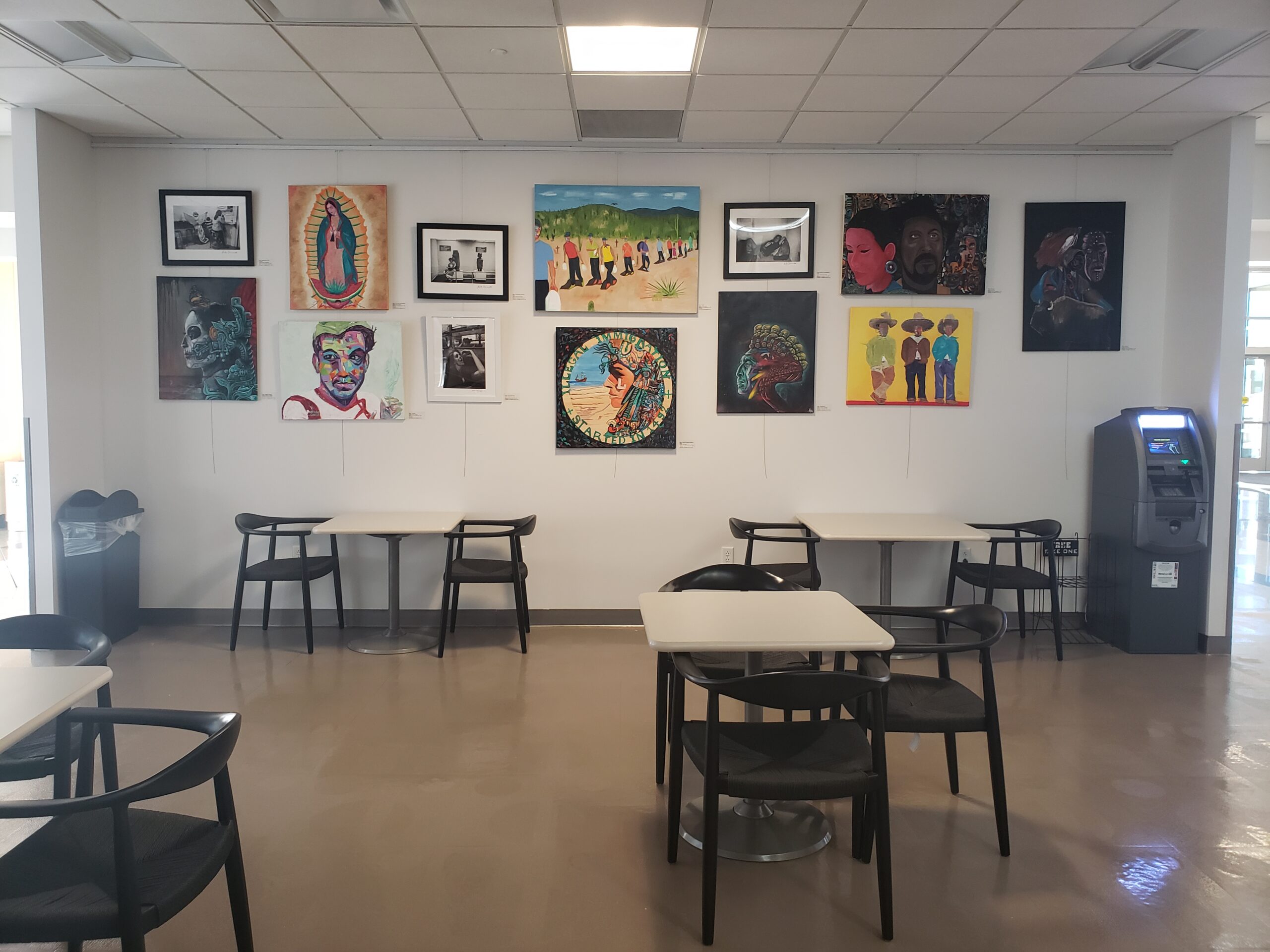 Art gallery in a cafeteria with diverse colorful paintings, tables, chairs, and an ATM.
