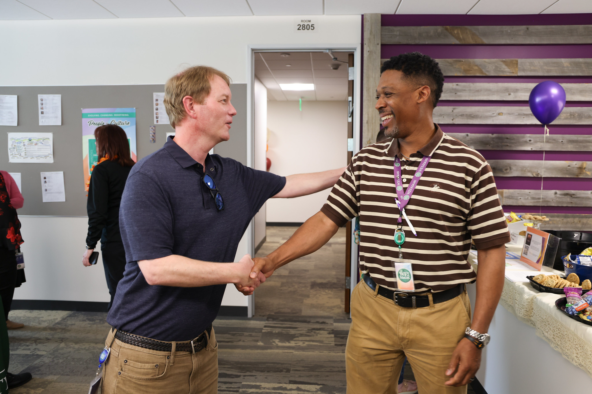 Two men shaking hands and smiling in an office setting labeled Room 2805, with snacks and balloons nearby.