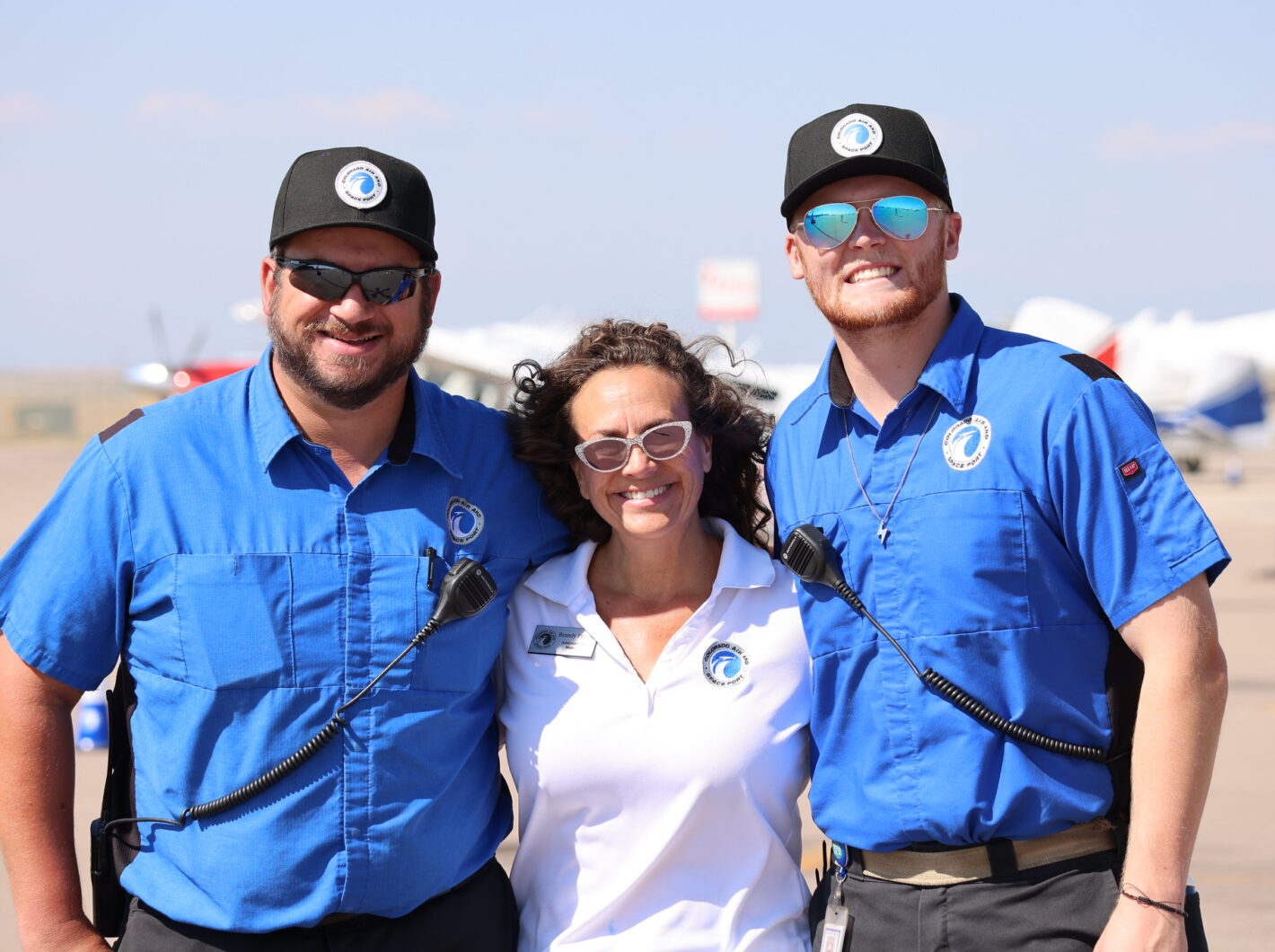 Three people smiling, wearing aviation uniforms with logos, standing on a sunny tarmac.