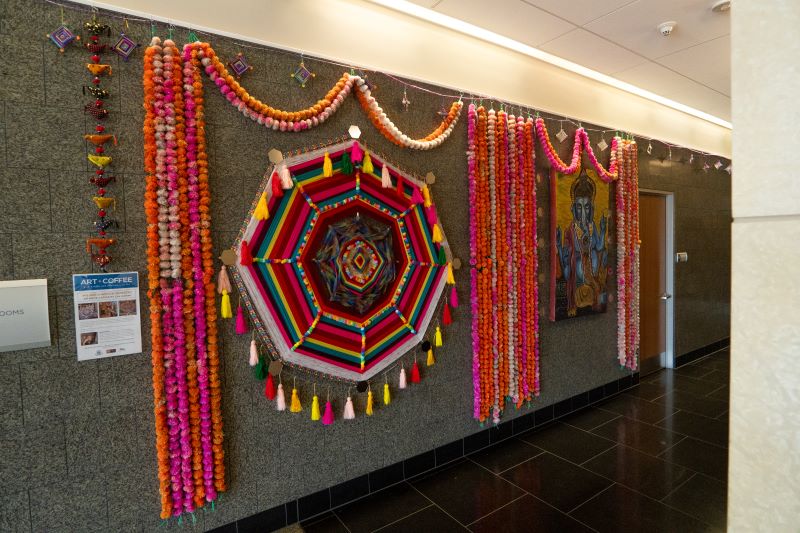 Colorful wall art display with vibrant floral garlands, geometric centerpiece, and framed painting on a gray wall.