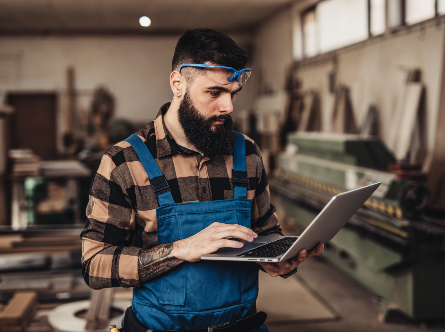Man in workshop using a laptop, wearing safety goggles and blue overalls.