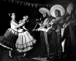 Two women in traditional dresses dance joyfully, accompanied by three musicians in sombreros playing instruments.