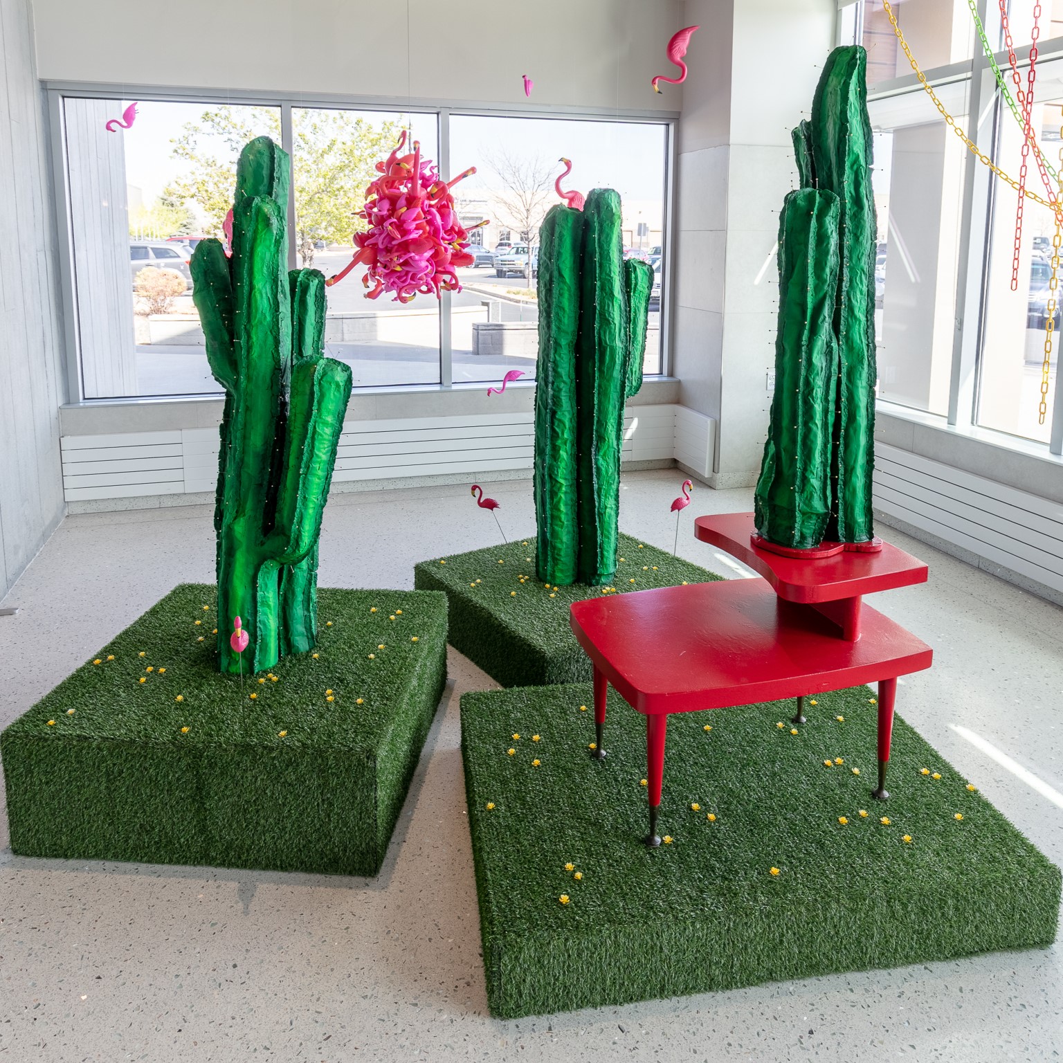 Colorful art installation with tall green cacti, pink flamingos, and a red table on artificial grass.