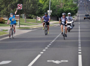 People biking on a road with a police motorcyclist nearby, one cyclist raising a hand, "No U-turn" sign visible.