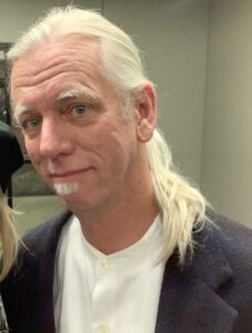 Craig Rouse with long white hair in a ponytail, wearing a white shirt and jacket, smiling gently