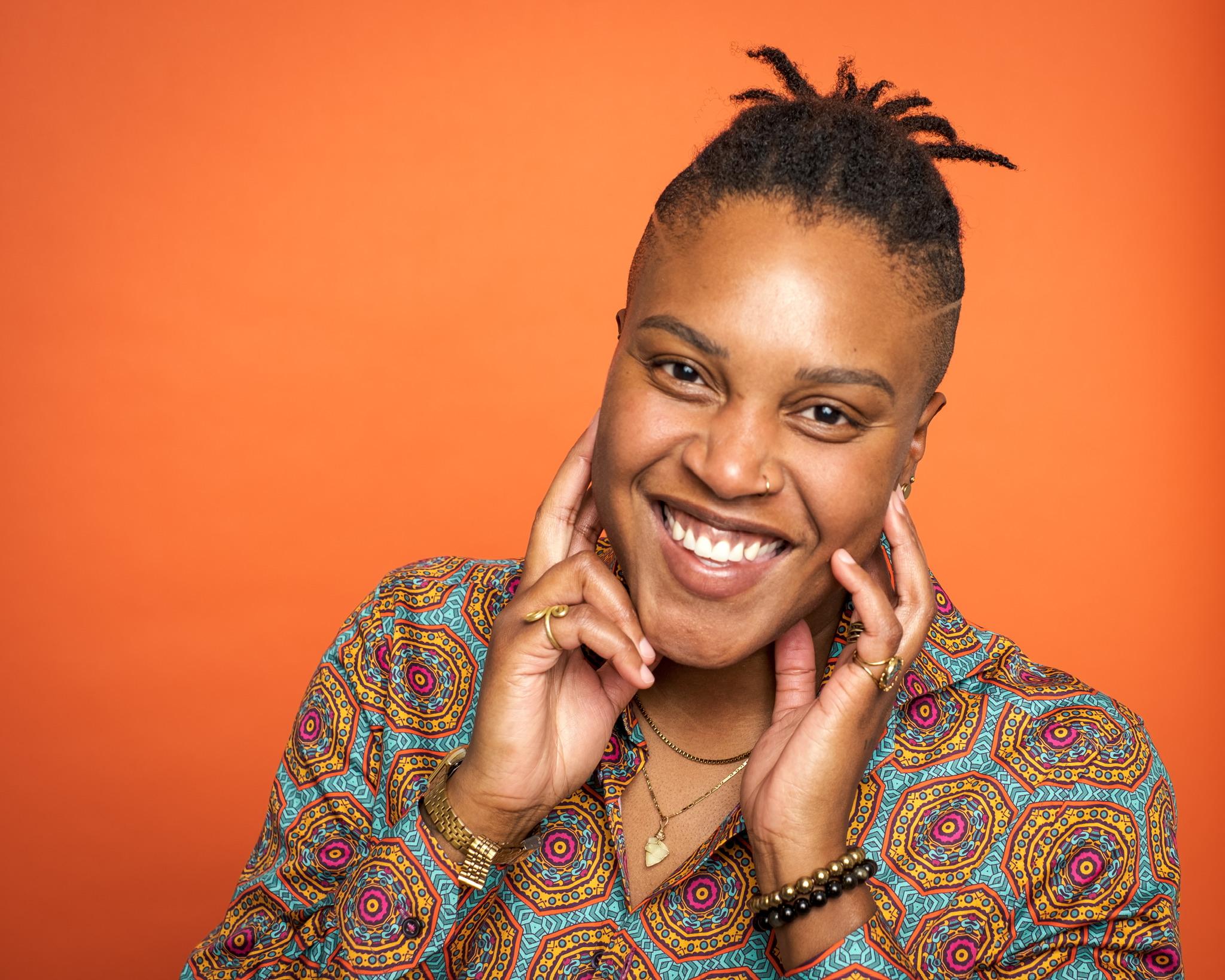 DSCF1192.jpg smiling person against orange backdrop, hands near face, wearing patterned shirt