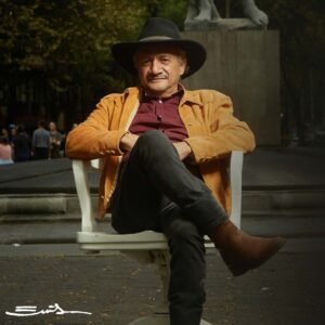 Emilio sits confidently outside in a black hat and brown jacket.