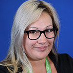 Gina Mora smiling, wearing glasses and a lanyard, blue background
