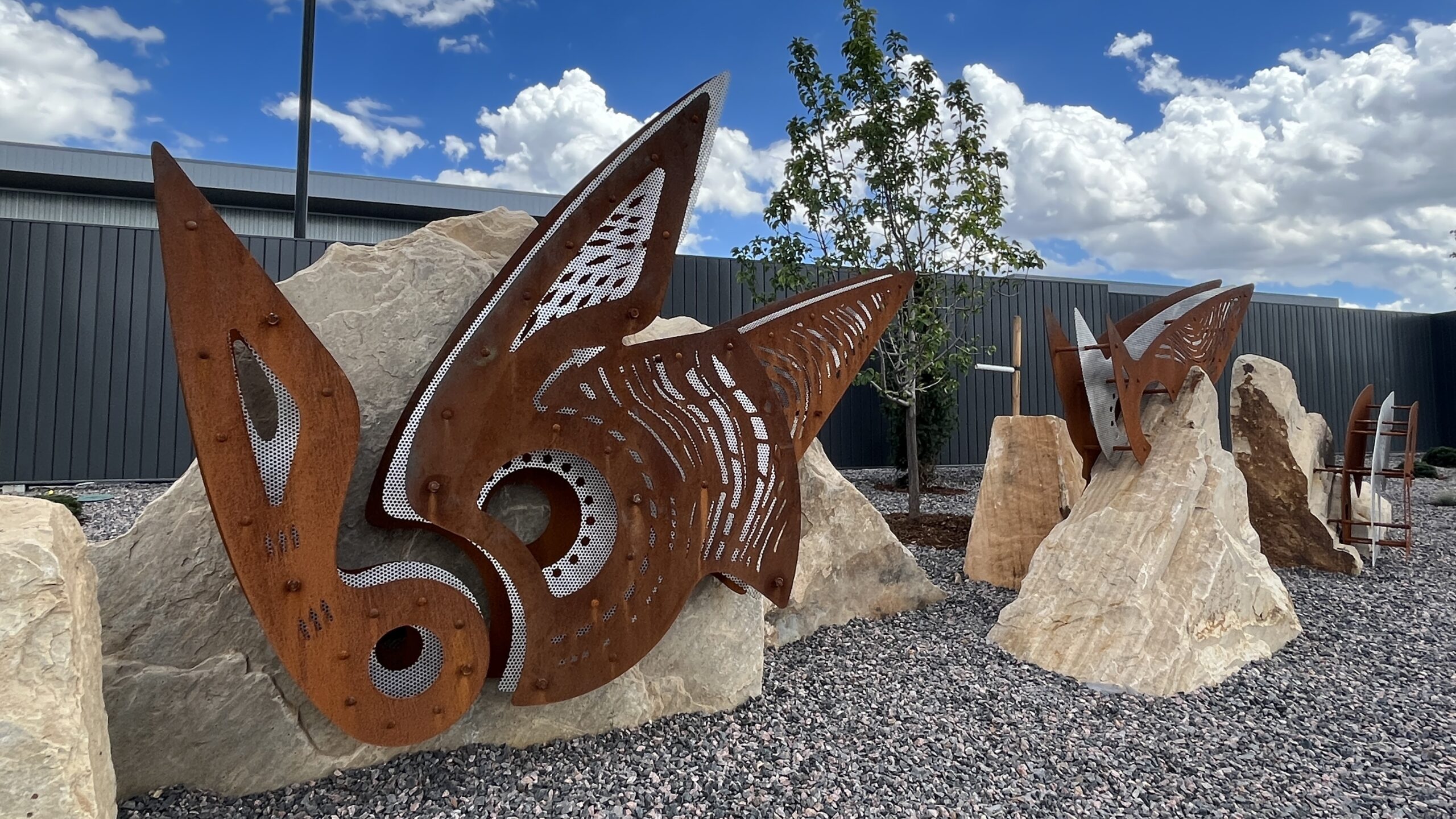 Rusty metal sculptures resembling butterfly wings embedded in large rocks, with a tree and cloudy sky backdrop
