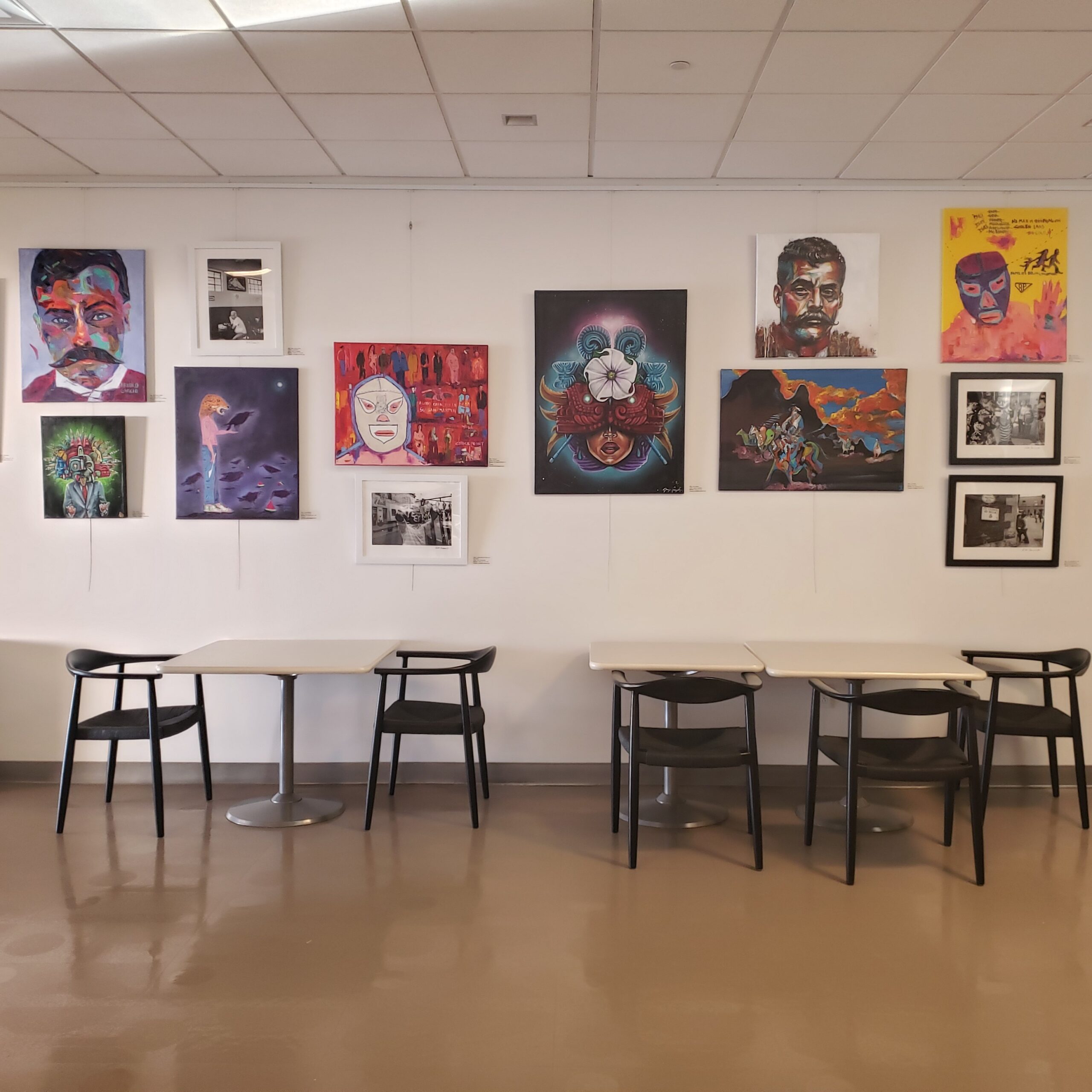 Art gallery wall with vibrant portraits and abstract paintings above tables and chairs.
