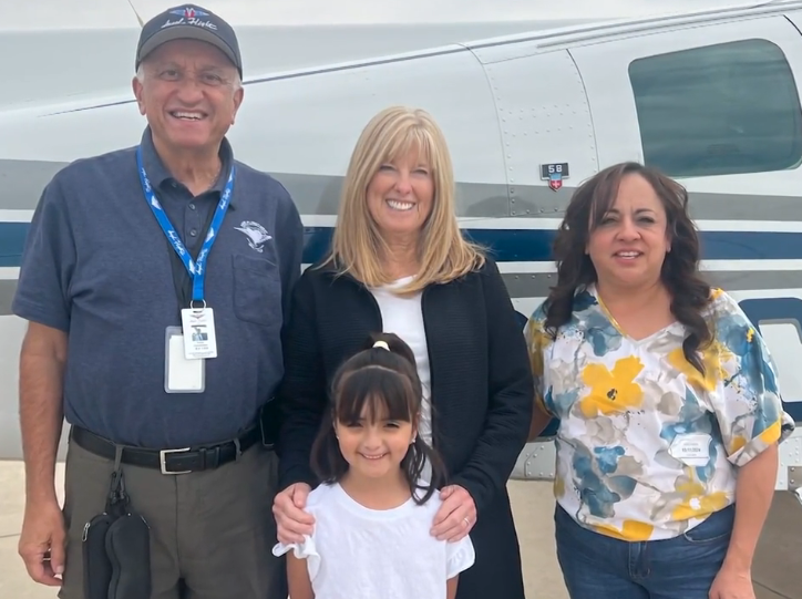 Itzy, Mirna, and Nancy smiling with two adults in front of airplane.