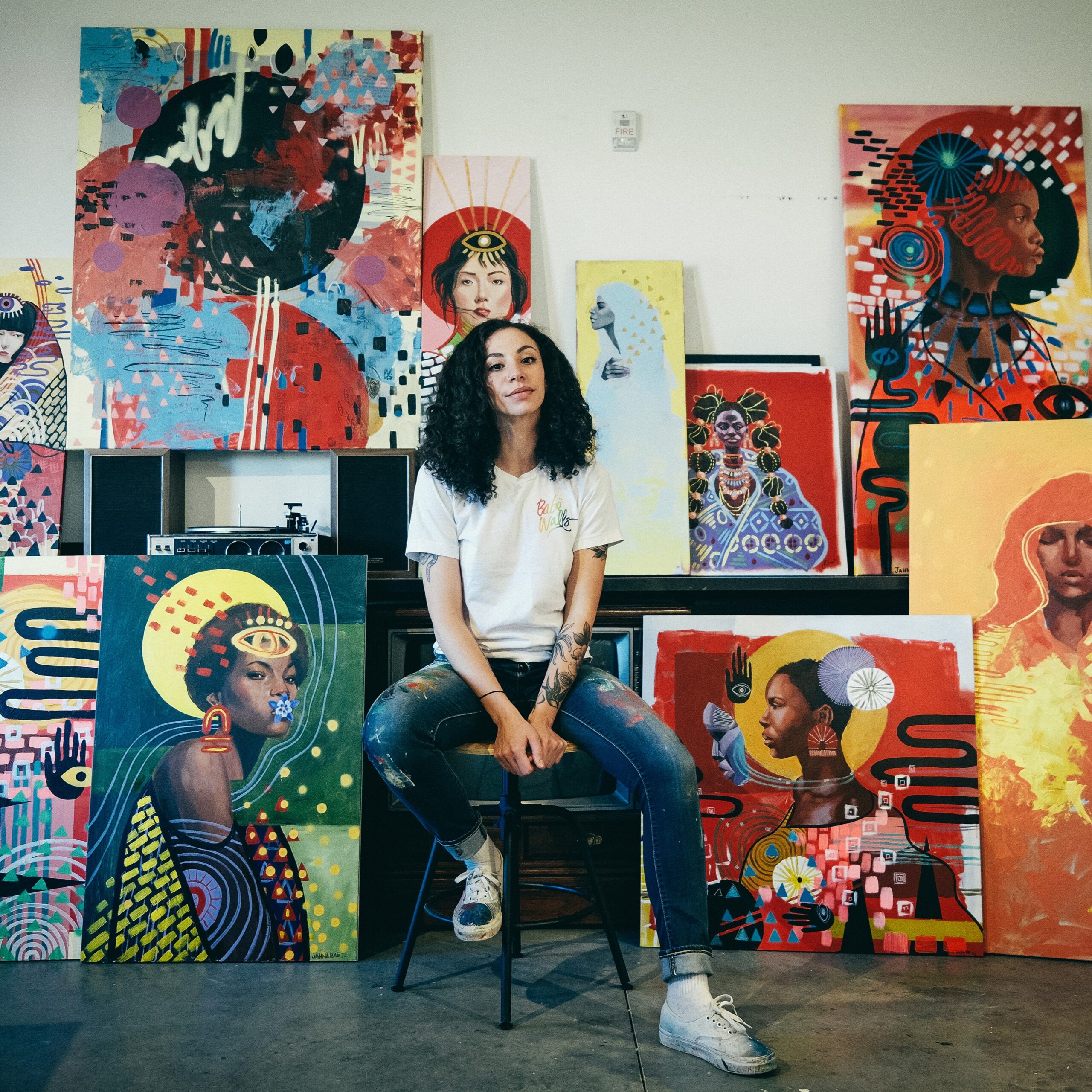 Jahna-Rae sits on a stool surrounded by vibrant, abstract paintings in a studio.
