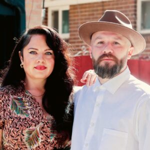 Jolt and CoCo embracing outdoors, both looking at camera. CoCo in patterned shirt, Jolt in white shirt with hat.