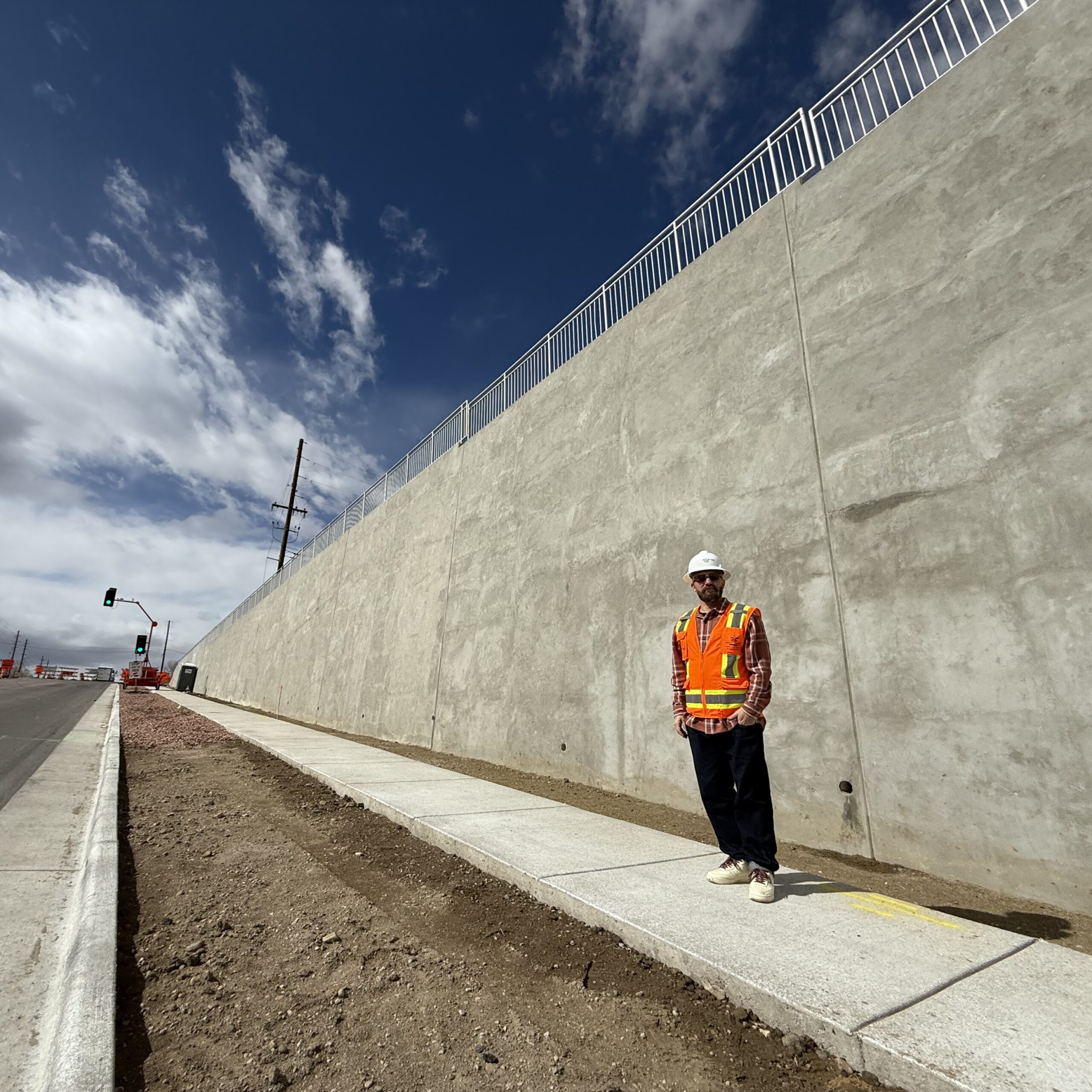Jolt standing on a sidewalk beside a tall concrete wall, wearing a safety vest and helmet, under a blue sky.