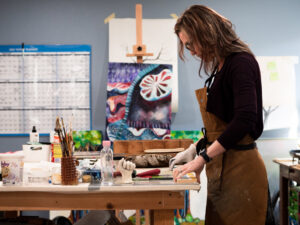 Melody-Headshot.jpg creates art in a studio, surrounded by brushes and colorful paintings.