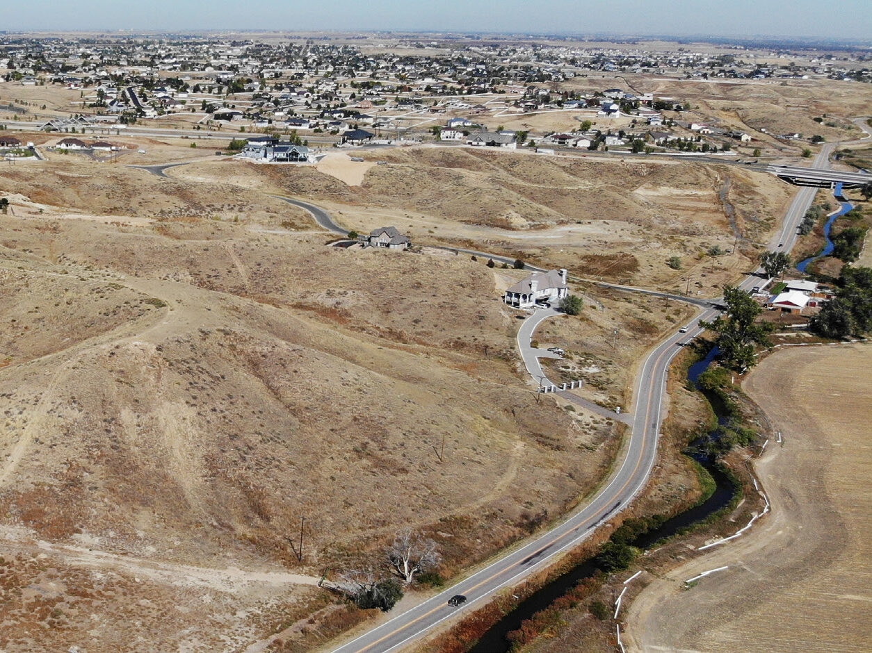 Aerial view of a dry, hilly landscape with scattered houses and winding roads leading to a distant suburban area.