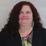 Shannon Lillie smiling with long curly hair, wearing earrings and a colorful shirt under a black jacket.
