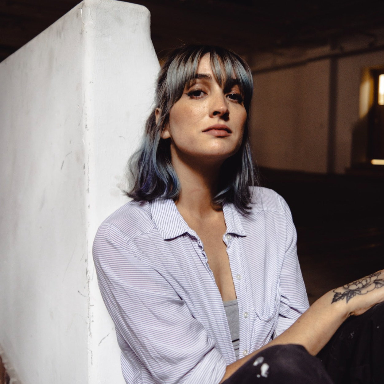 Sidney Masuga leaning against a white wall, wearing a striped shirt, looking serene.
