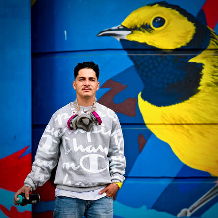 Person standing confidently in front of colorful bird mural, wearing casual clothes and a respirator around neck.