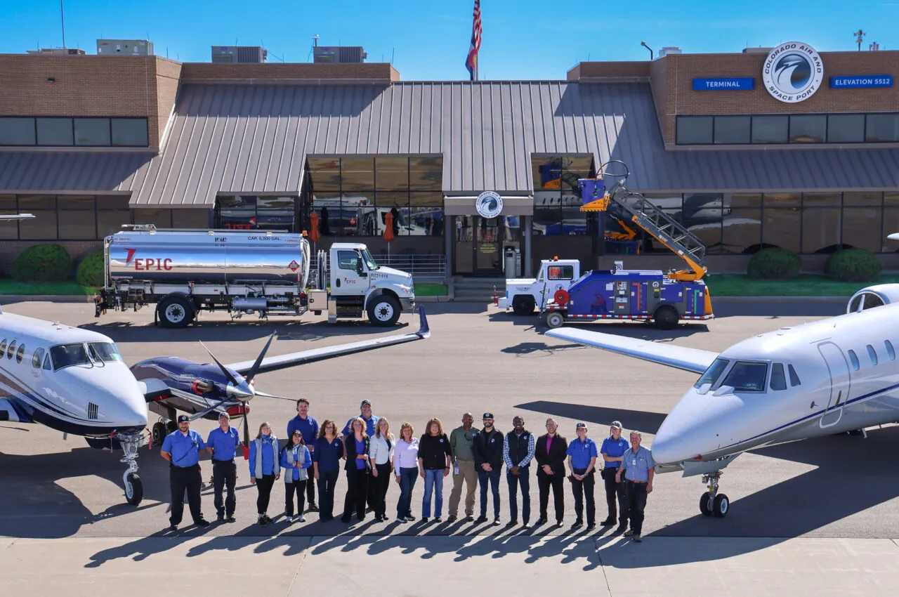 People standing in front of two planes at Colorado Air and Space Port terminal, with service vehicles in background