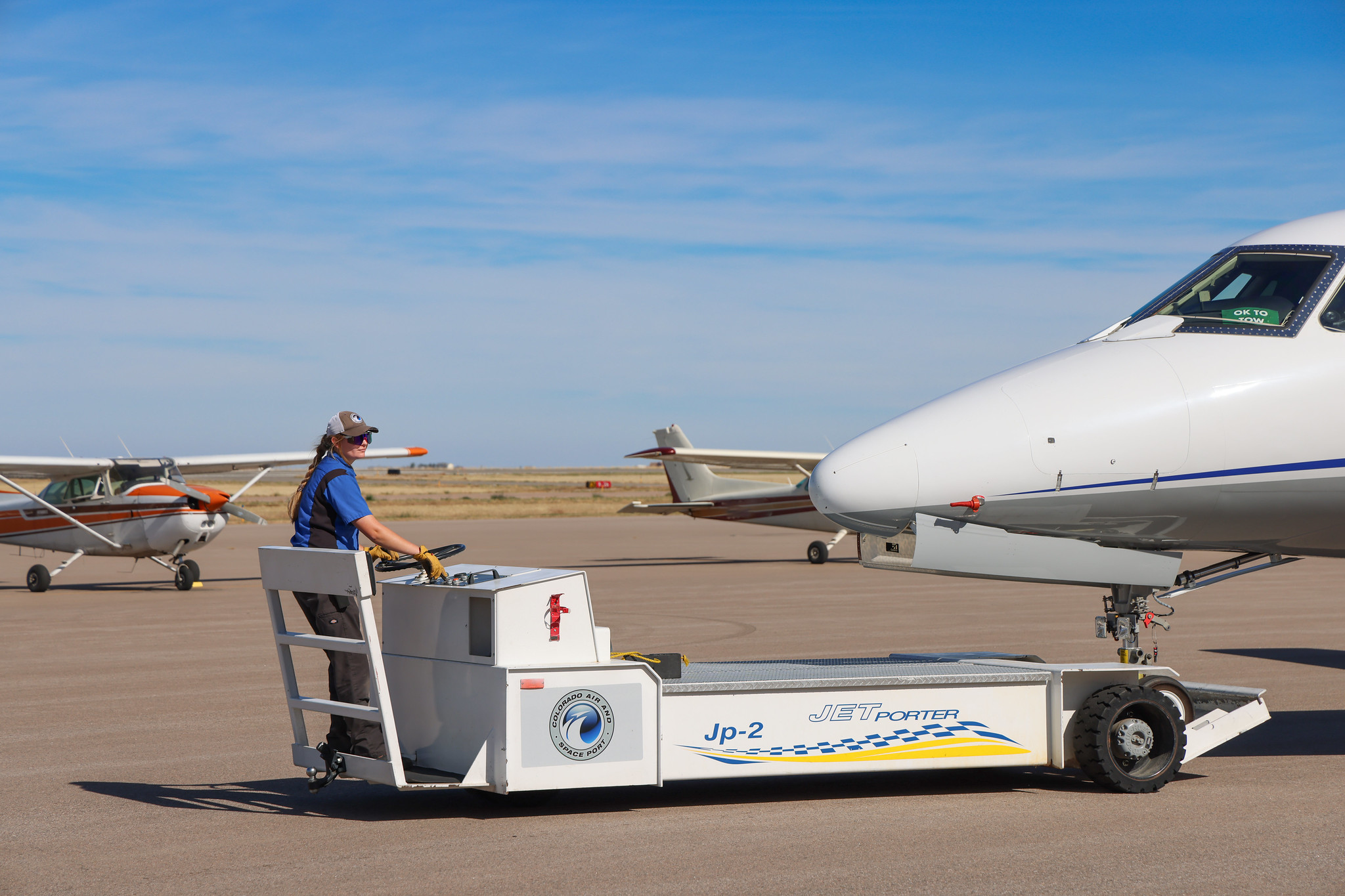 Person operating aircraft towing vehicle at airport, with two small planes parked in the background.