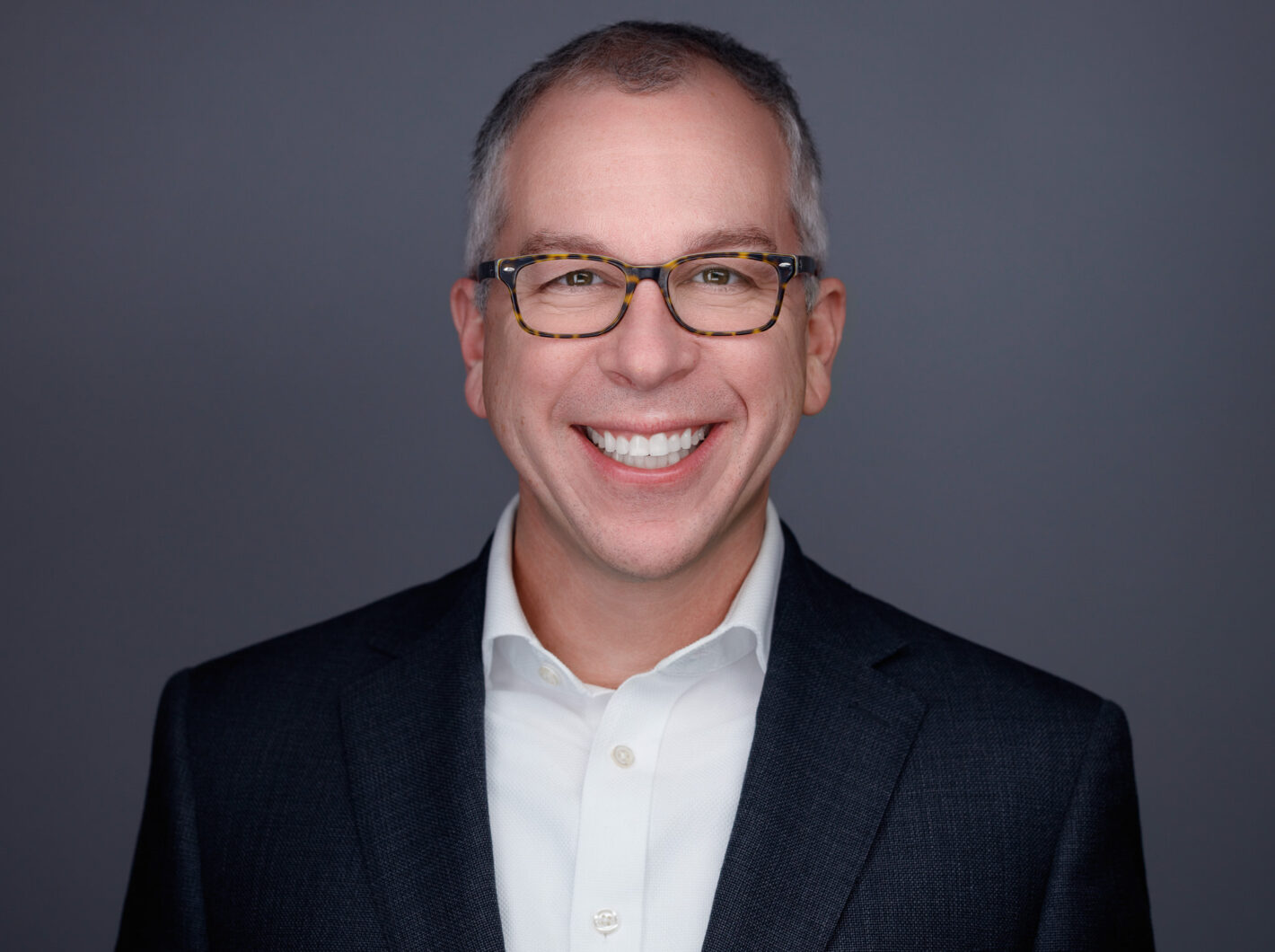 Jason Schultz smiling in a suit and glasses against a gray background