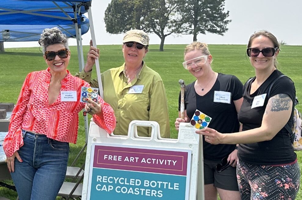 Four smiling women stand outdoors, holding coasters beside a sign reading, "Free Art Activity: Recycled Bottle Cap Coasters."