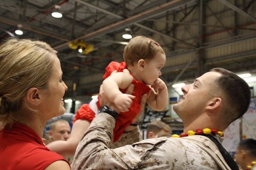 Family4_2.jpg features a soldier joyfully lifting a baby girl in a red outfit. A woman smiles beside them in a hangar setting.