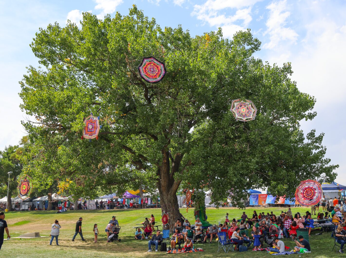 Crowd sitting under a large tree with colorful, patterned woven decorations hanging. Nearby tents indicate a festival or market atmosphere.