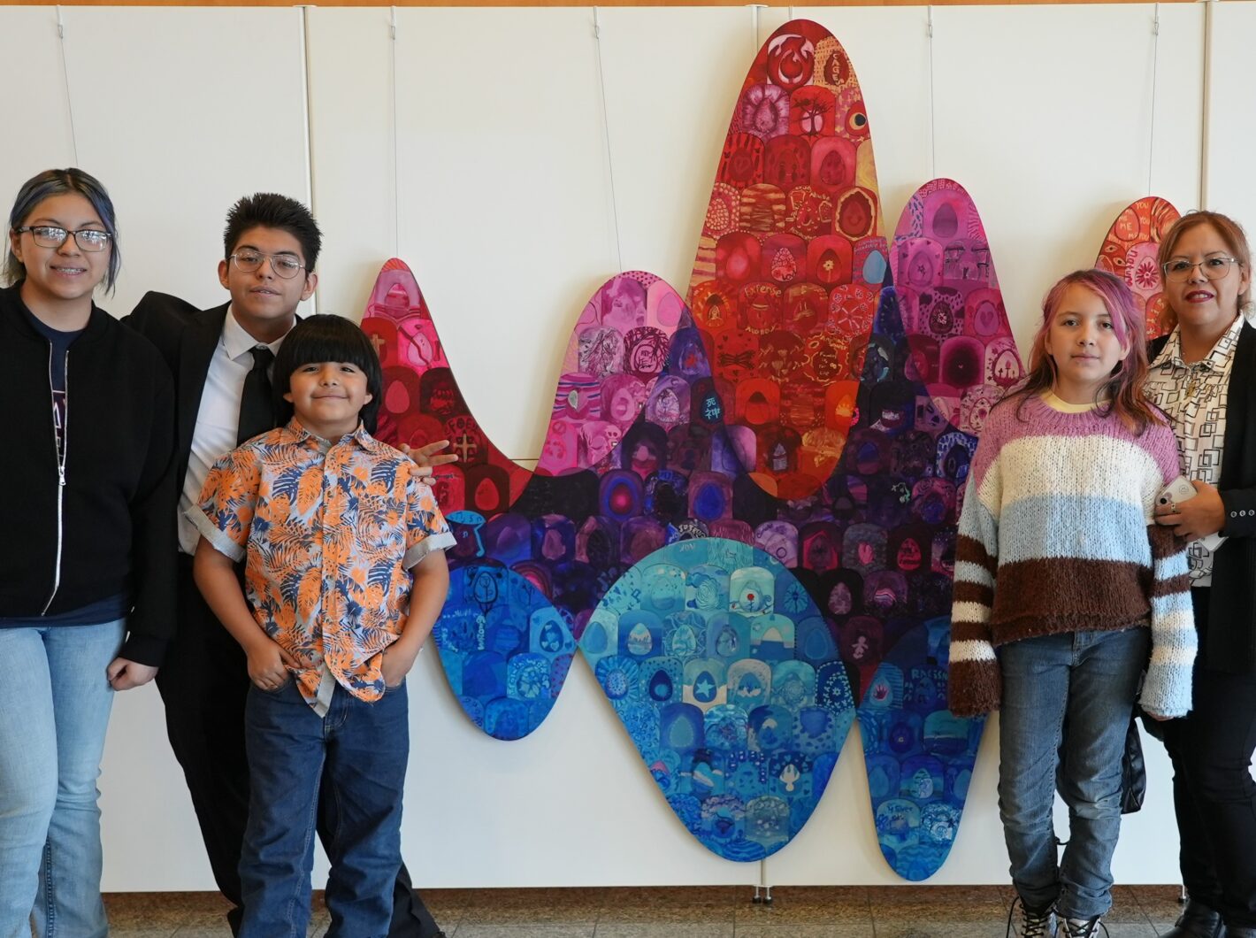 Five smiling people standing in front of colorful wave-shaped artwork, featuring reds, purples, and blues, on a white background.