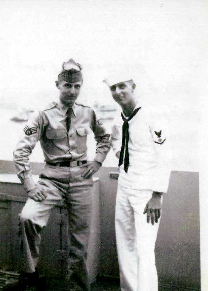 Two men in military uniforms standing together, one in a khaki uniform with sergeant stripes, the other in a white sailor uniform with an eagle insignia.