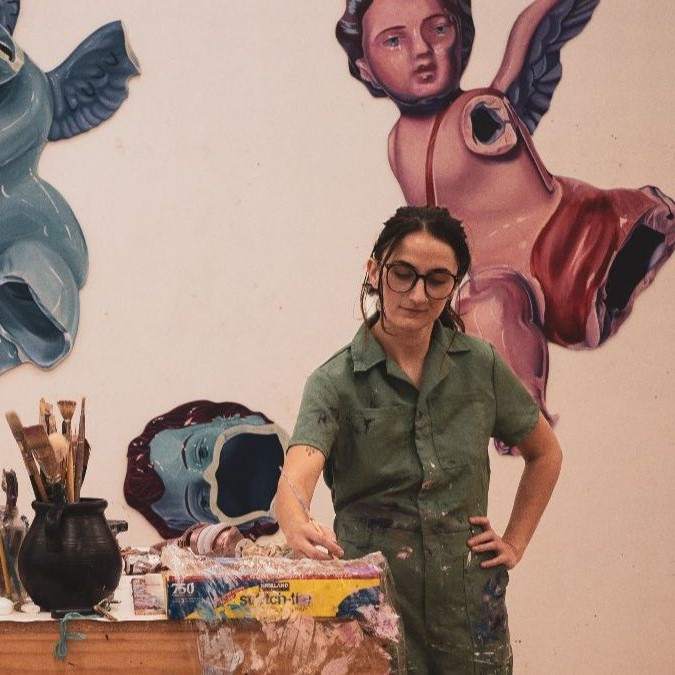 Jenna painting with brushes and tools, colorful angel artworks on wall behind.