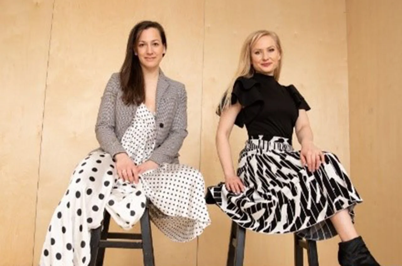 Two women sitting on stools, wearing polka dot and patterned skirts, smiling confidently.