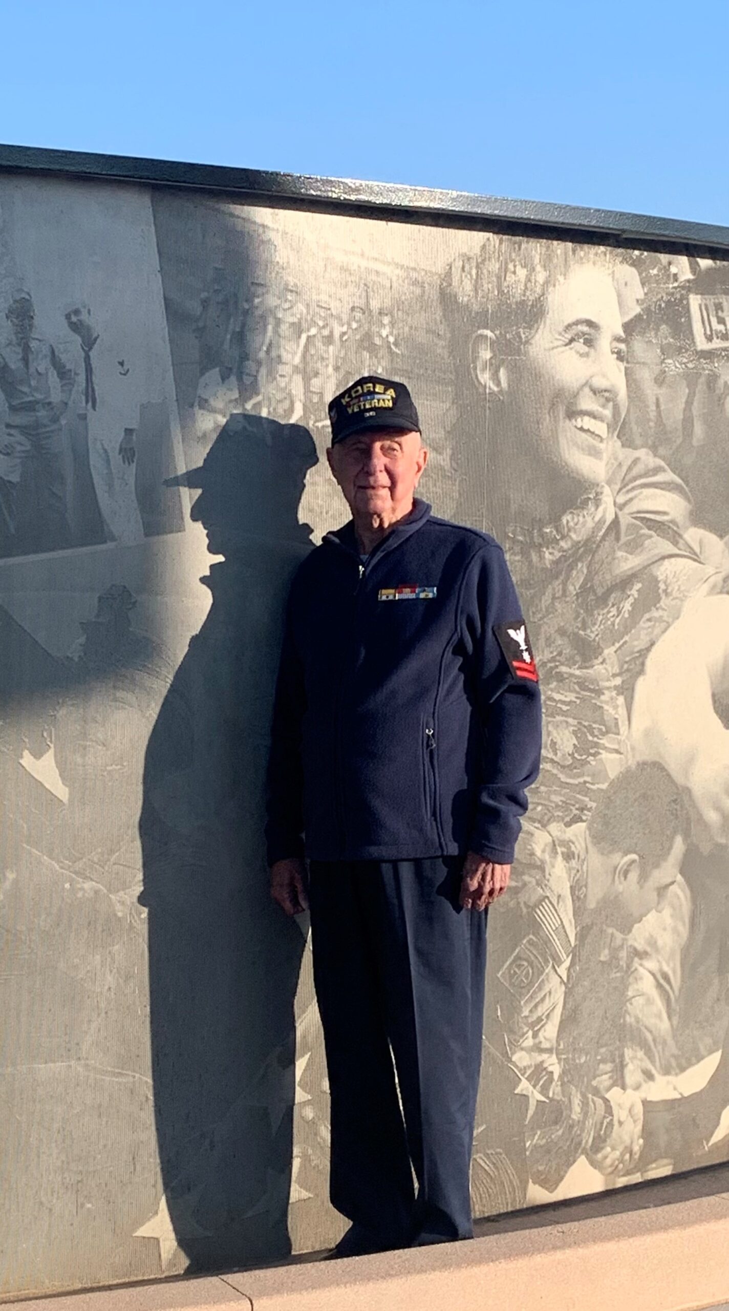 Peggys-Dad standing proudly in front of a Korean War memorial wall, wearing a "Korea Veteran" hat.