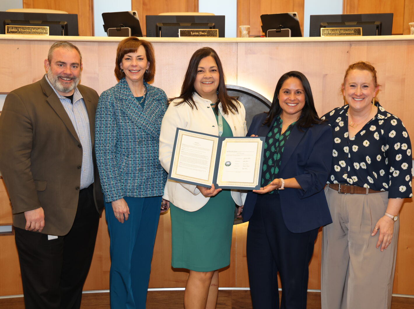 Five people standing together, two holding a framed certificate, smiling in a formal setting.
