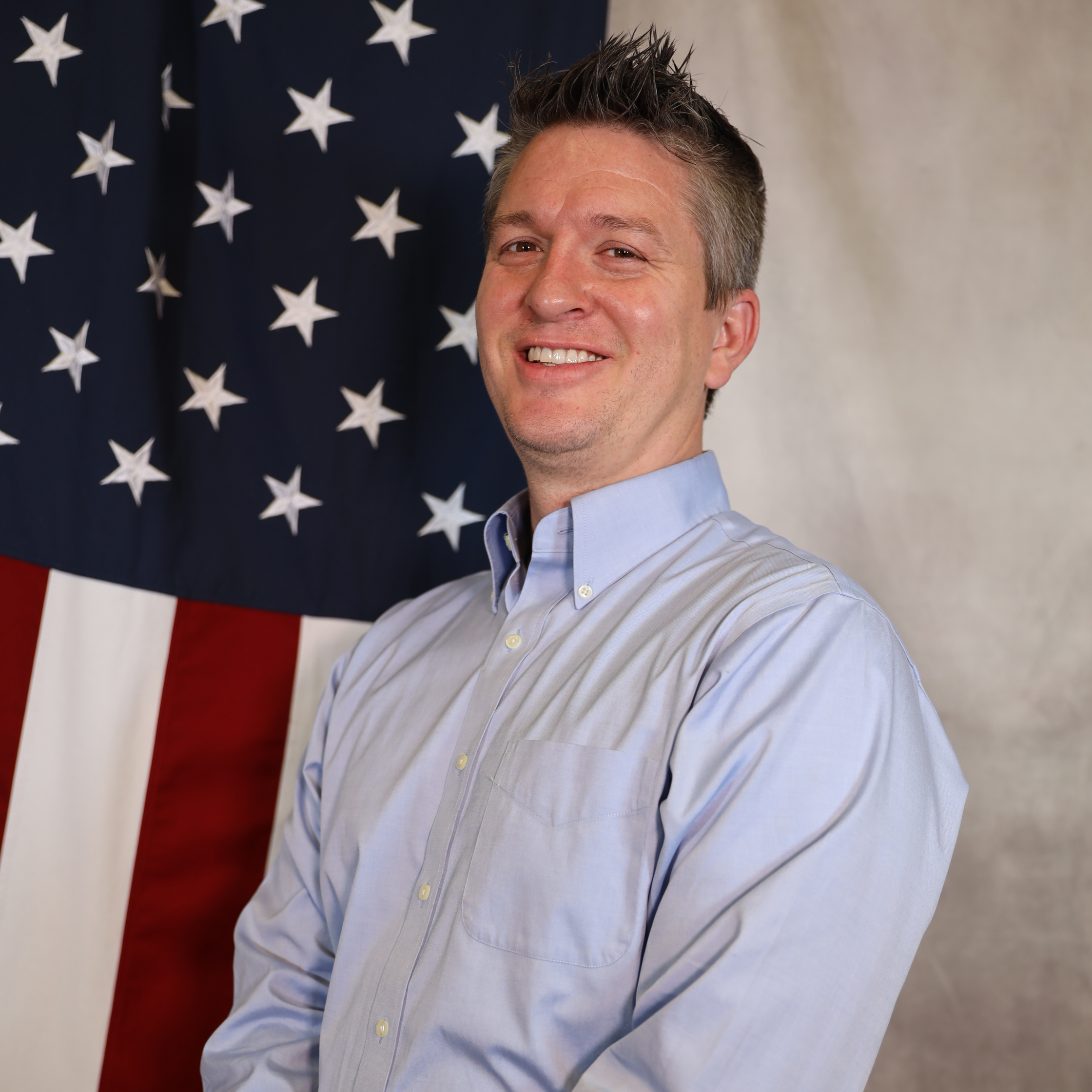 Bean smiling with American flag backdrop, wearing a light blue shirt.