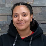Mia McKee smiling, wearing a black hoodie, with a stone wall background