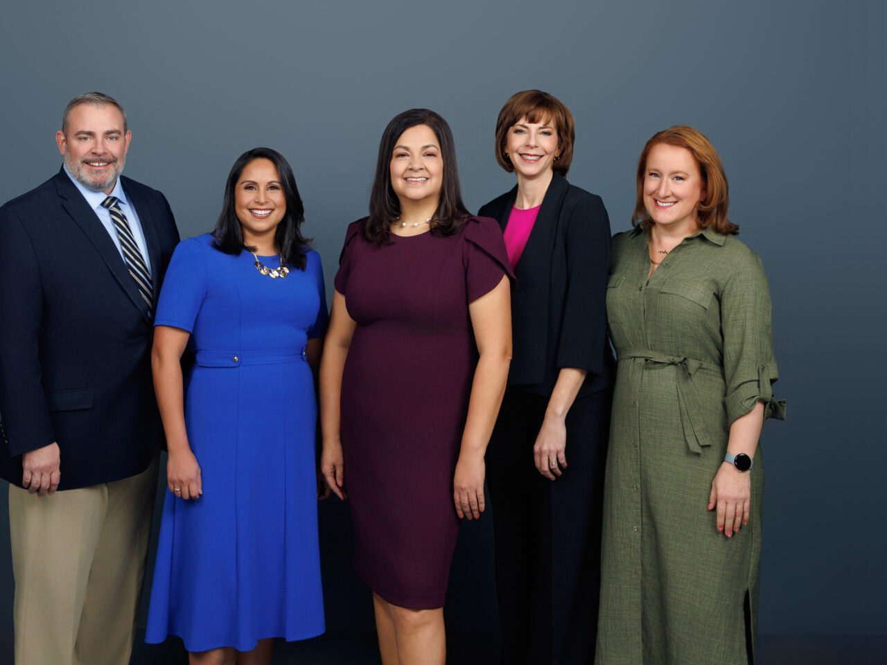 Five smiling people standing together against a gray background, dressed in formal attire