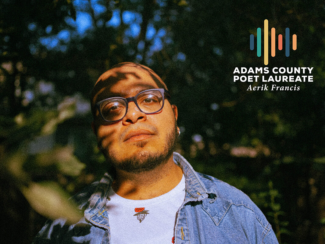 Aerik Francis standing outdoors, dappled sunlight, wearing glasses and denim jacket. Text: "Adams County Poet Laureate, Aerik Francis" with colorful logo bars.