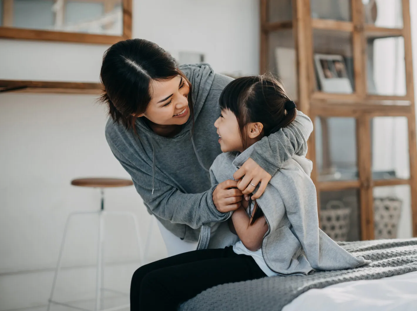 Mother in gray hoodie lovingly embraces smiling daughter in matching outfit, sitting indoors.