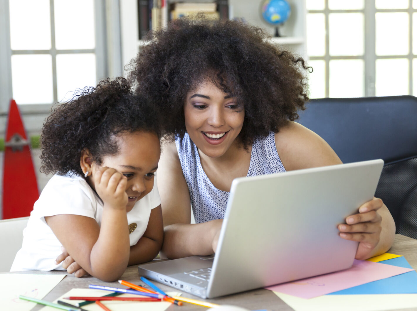 Child and adult smiling at a laptop on a desk with colorful papers and pencils.