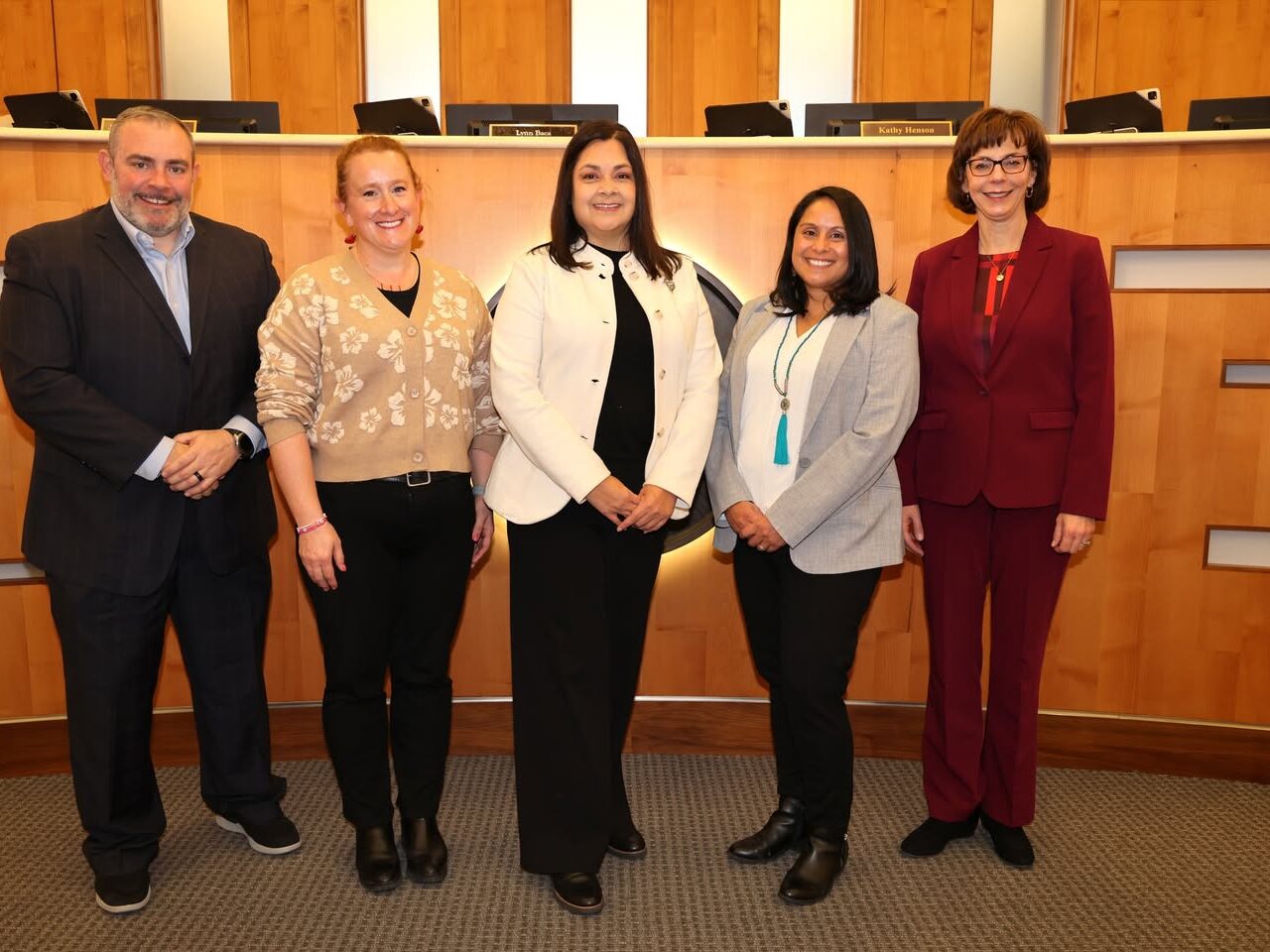Five people standing professionally in front of a wooden council chamber backdrop, smiling, with a plaque in view.