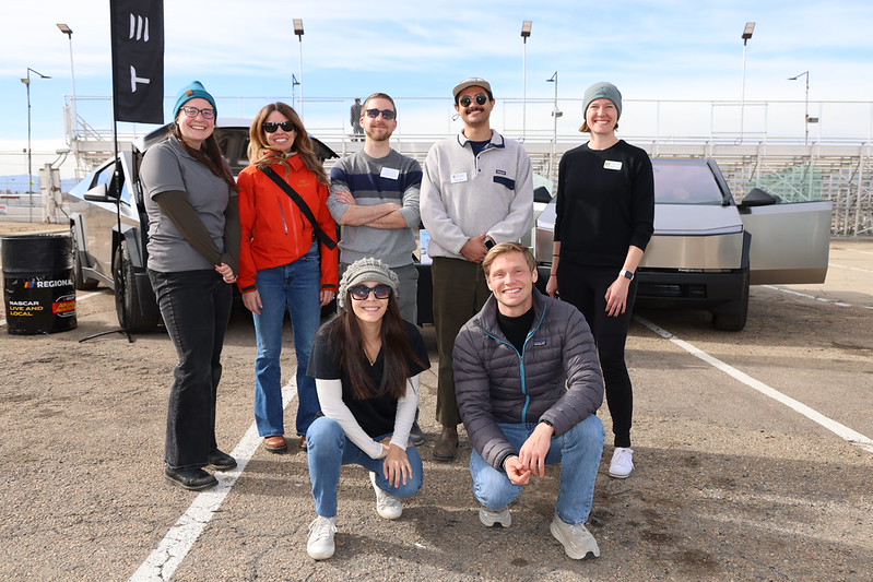 Group of eight people smiling outside near a vehicle, sunny day