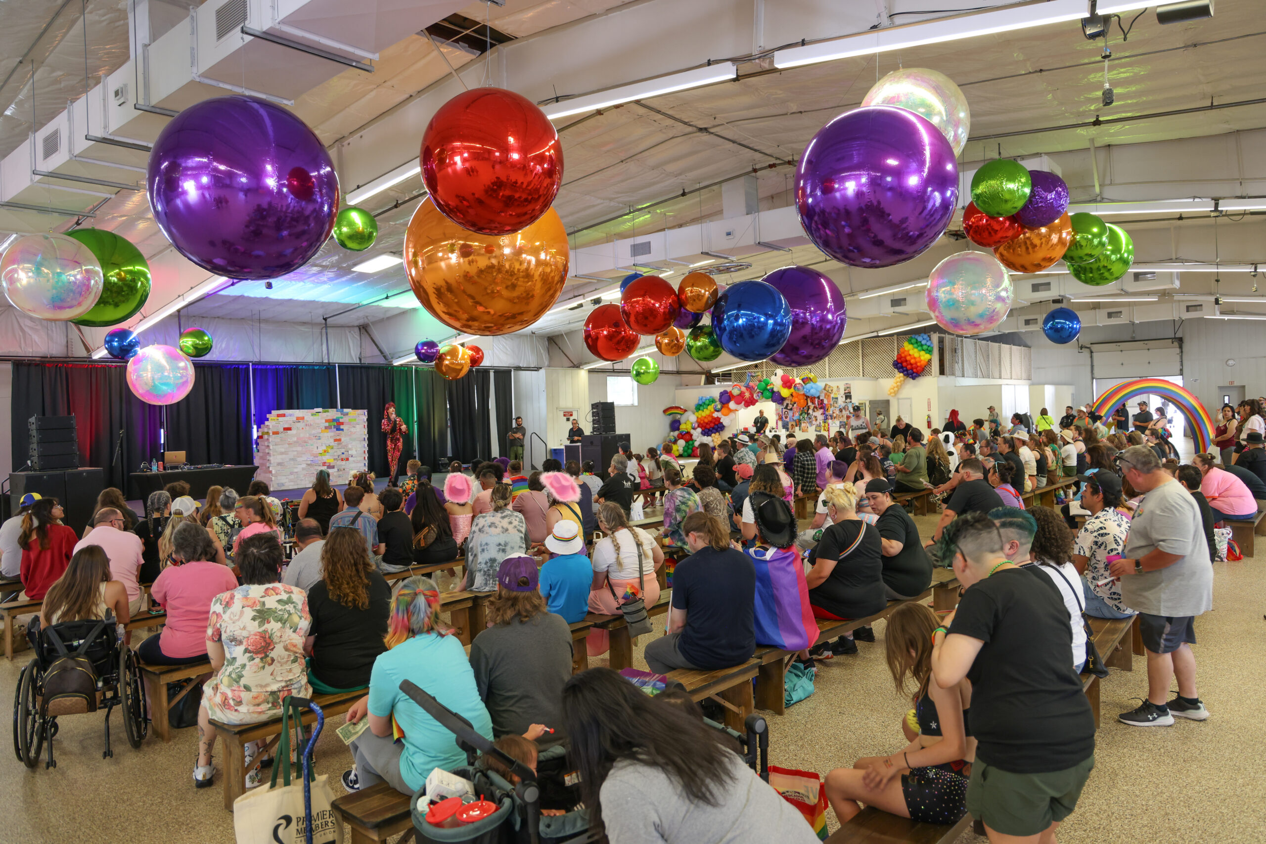 Colorful indoor Pride event with audience watching a performer on stage; large rainbow balloons decorate the ceiling