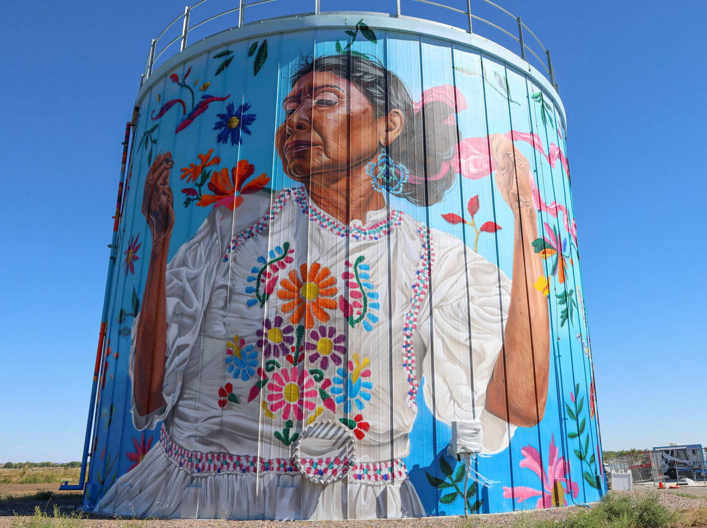 Mural of a woman in traditional dress with vibrant floral embroidery on a large cylindrical structure against a blue sky
