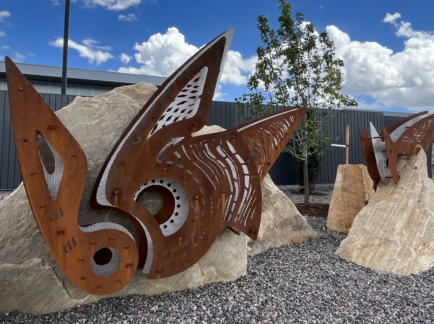 Rusty metal sculptures resembling butterfly wings embedded in large rocks, with a tree and cloudy sky backdrop