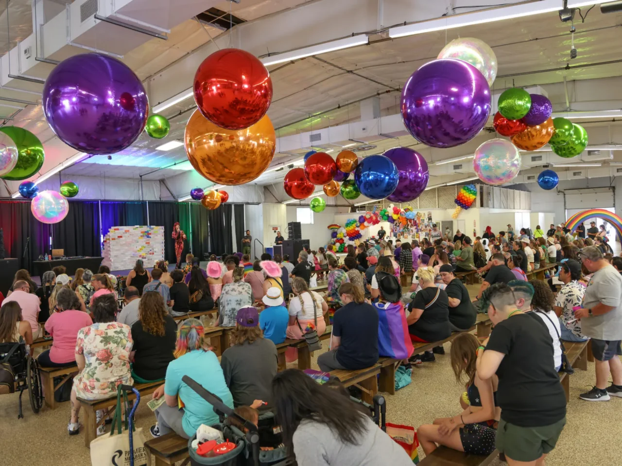 Colorful balloons hang over a lively indoor Pride event with a seated audience