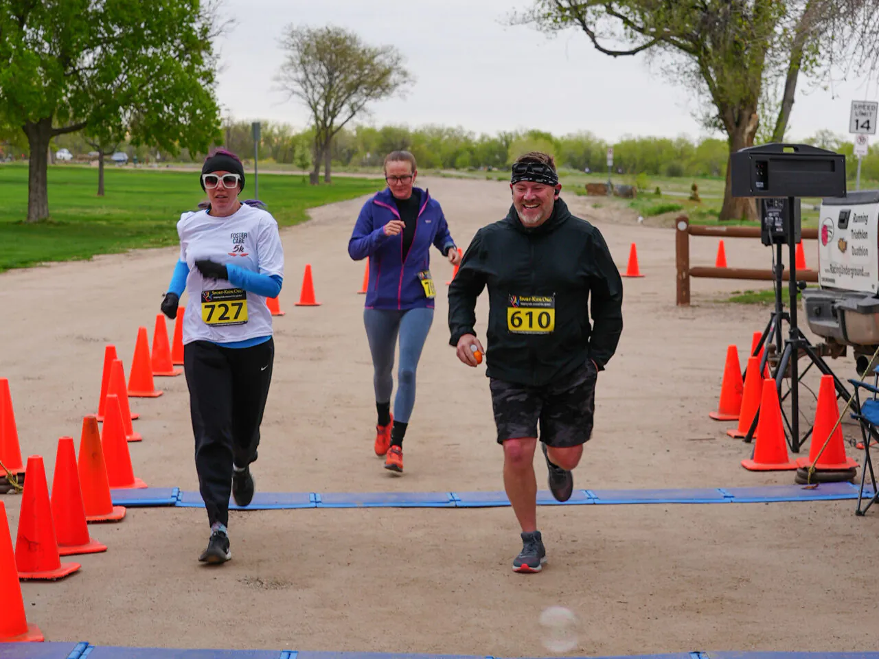 Three participants running near the finish line of a Foster Care 5K event, numbered vests 727 and 610, and orange traffic cones marking the path